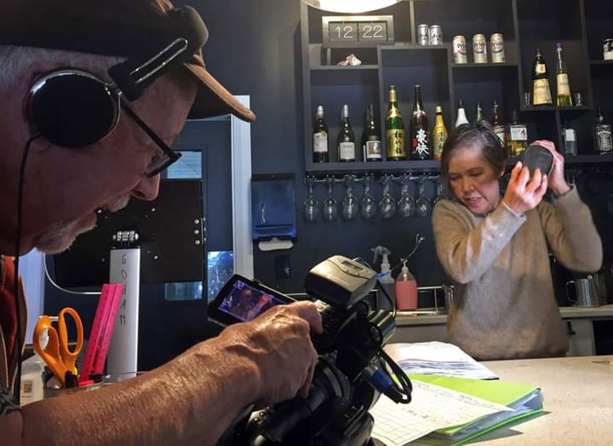 A woman shakes a cocktail behind a bar while a filmmaker films